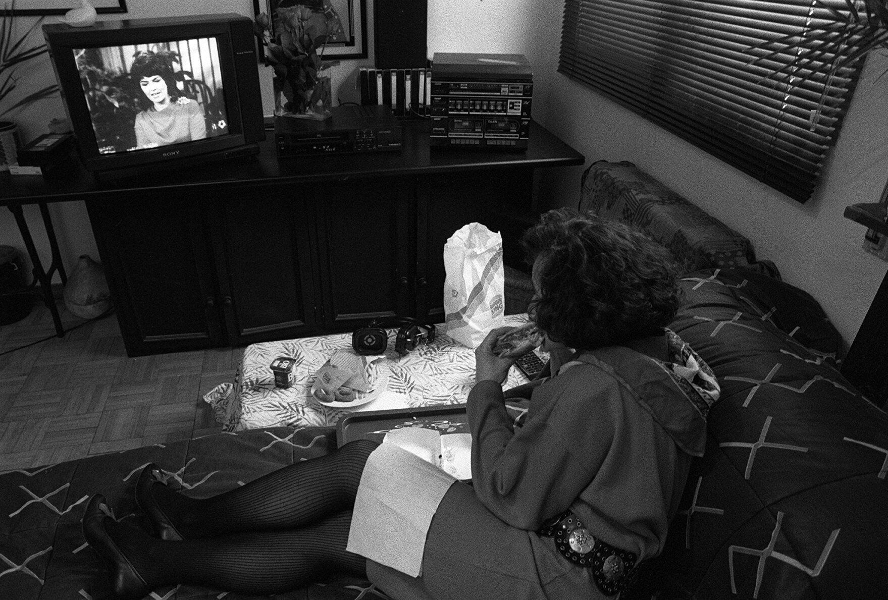Una mujer se come una hamburguesa mientras ve la televisión en su casa en 1990.