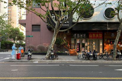 Unas personas en traje protector aguardan junto a unas tiendas cerradas por el confinamiento en Shanghái