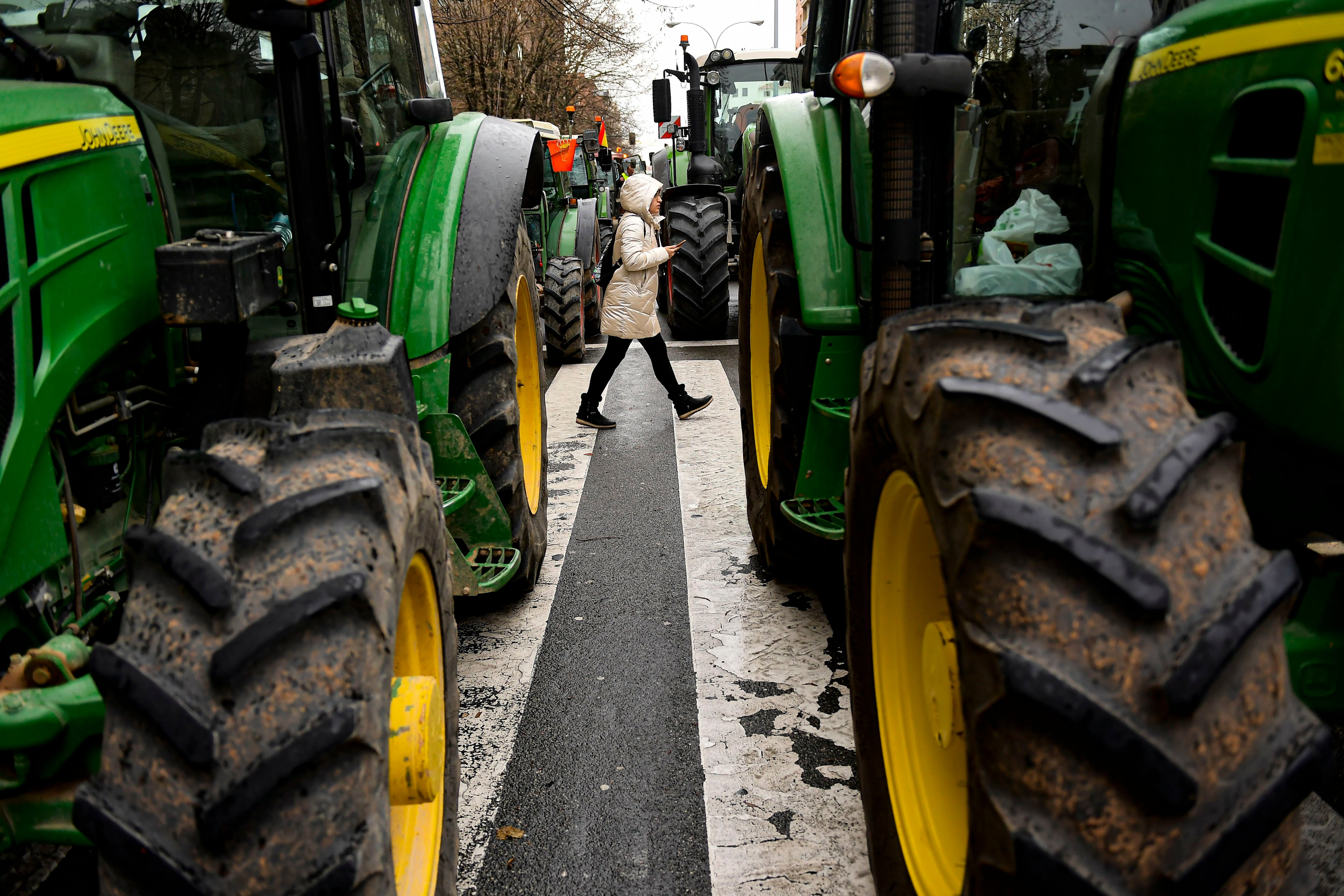 A pedestrian crosses a street between tractors in the center of the city while taking part in a protest, in Pamplona, northern Spain, Thursday, Feb. 8, 2024. For three non stop days farmers across Spain have staged tractor protests across the country, blocking highways and causing traffic jams to demand  changes in European Union policies and funds and measures to combat production cost hikes. (AP Photo/Alvaro Barrientos)