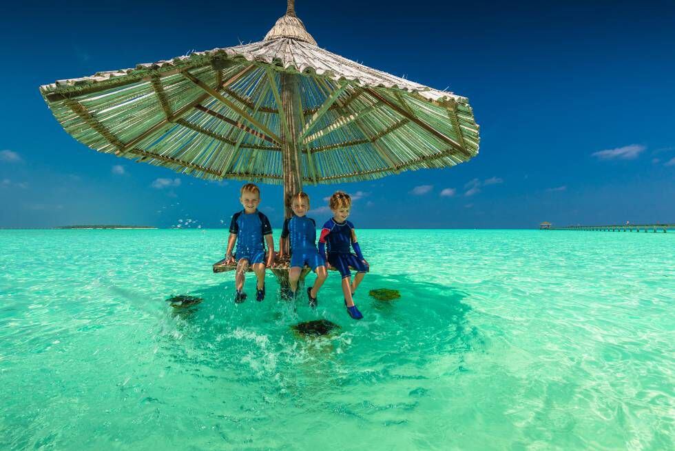 Tres niños sonríen debajo de una sombrilla clavada sobre la arena en mitad del mar.