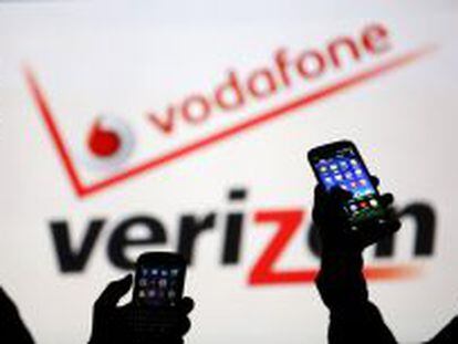 Dos usuarios miran sus m&oacute;viles ante los logotipos de Vodafone y Verizon. 