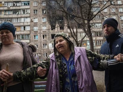 People help an elderly woman to walk in a street with an apartment building hit by shelling in the background in Mariupol, Ukraine, Monday, March 7, 2022. (AP Photo/Evgeniy Maloletka)