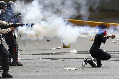 Una manifestante se arrodilla sosteniendo una piedra, mientras miembros de la policía Nacional venezolana dispara gases lacrimógenos.