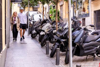 Dos jóvenes pasean por una acera estrecha del centro de Madrid, entre coches y motos aparcados, en una calle de plataforma única. 