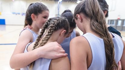 A group of high school girls huddles together with their coach before a basketball game. Their arms are around each other.