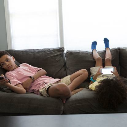 Dos niños están en el sofá. Uno mira la televisión y el otro juego su teléfono.