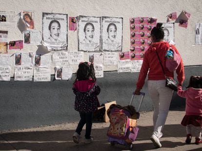 Una mujer camina con dos niñas frente a varios carteles de mujeres desaparecidas.
