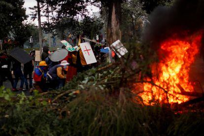 Un árbol derribado y en llamas en primer plano, mientras manifestantes utilizan escudos improvisados para protegerse.