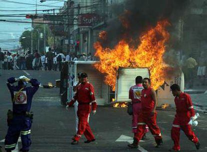 Miembros de los servicios de emergencia observan un vehículo de la televisión incendiado en los disturbios del sábado en San Salvador.