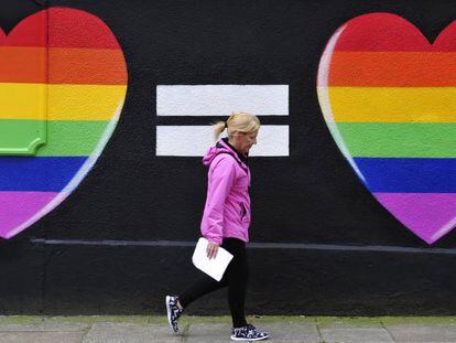 Una mujer camina junto a un muro pintado con dos corazones con los colores del arcoíris, en Dublín.