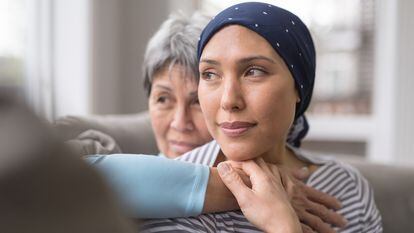An ethnic woman wearing a headscarf and fighting cancer sits on the couch with her mother. She is in the foreground and her mom is behind her, with her arm wrapped around in an embrace, and they're both looking out the window in a quiet moment of contemplation.