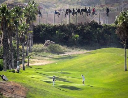 Una docena de inmigrantes permanece encaramado a la valla de Melilla mientras dos personas juegan al golf.