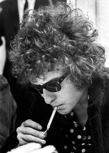 Dylan durante una conferencia de prensa en Suecia (1966). Convertido al rock, y siendo parte de la Generación Beat, su imagen evoluciona dejándose crecer el cabello –que rizado y revuelto le da un aire sexy–; utiliza las Way Farer, de Ray Ban y comienza a vestirse de riguroso negro con camisas estampadas que se erigen como icono rockero. Mención especial a la longitiud de las uñas de la mano derecha con la que se enciende el cigarrillo.