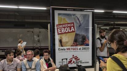 Unos turistas esperan un tren en la estación de Barcelona Sants junto a un cartel de lucha contra el ébola en 2014.