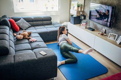 Una madre hace ejercicio frente al televisor mientras su bebé descansa en el sofá.