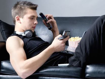 Un joven tumbado en un sofá utilizando un teléfono móvil, un ordenador portátil y un MP4.