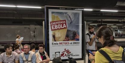 Unos turistas esperan un tren en la estación de Barcelona Sants junto a un cartel de lucha contra el ébola en 2014.