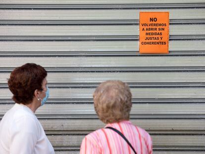 Dos mujeres leen un cartel puesto en una persiana echada de un comercio de Málaga en el que se puede leer: "No volveremos a abrir sin medidas justas y coherentes", este viernes.