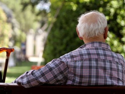 Un anciano de espaldas sentado en un banco junto a su bastón. 