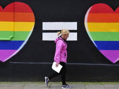 Una mujer camina junto a un muro pintado con dos corazones con los colores del arcoíris, en Dublín.