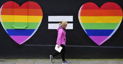 Una mujer camina junto a un muro pintado con dos corazones con los colores del arcoíris, en Dublín.