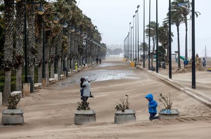 Una mujer cruza el paseo marítimo de Valencia cubierto por la arena durante el termporal de lluvia y viento.