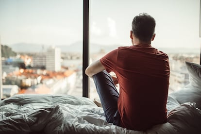 Un hombre sentado en la cama mira por la ventana.