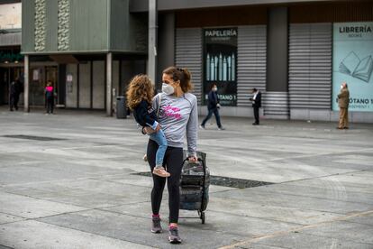 Una madre con mascarilla camina a casa con su hija en brazos después de hacer la compra, en la plaza de Felipe II (Madrid).