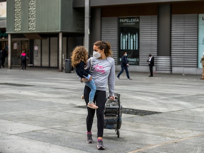 Una madre con mascarilla camina a casa con su hija en brazos después de hacer la compra, en la plaza de Felipe II (Madrid).