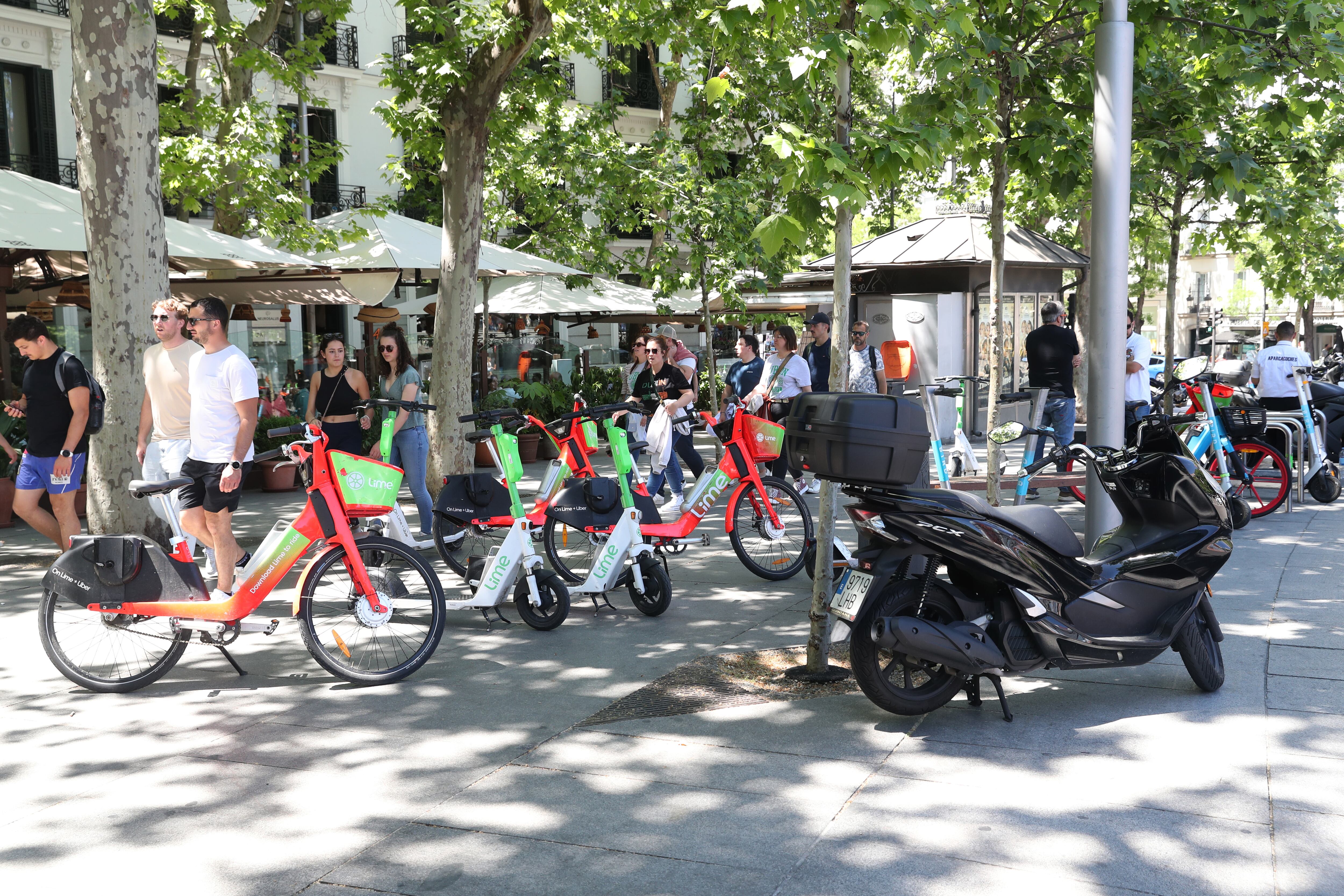 Patinetes, bicicletas y motos aparcados en una acera del centro de Madrid; al fondo, varias terrazas. 