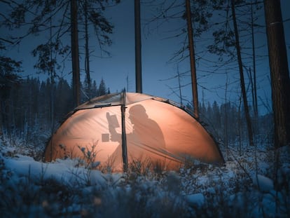 Un hombre lee un libro dentro de una tienda de campaña en mitad de un bosque nevado.