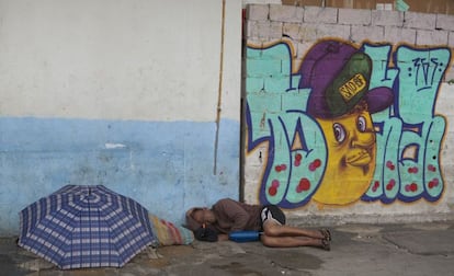 Homem dorme com sua garrafa térmica na parte seca da calçada, em frente a um muro com grafite. Ao seu lado há outra pessoa, que também dorme protegida com um guarda-chuva e uma manta.