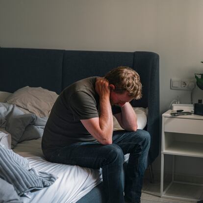 A depressed mature male sits on his bed. He clasps his hand behind his head