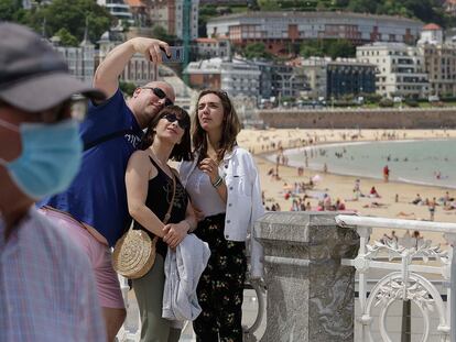 Un señor con mascarilla pasa por delante de un grupo de turistas que se fotografían en la playa donostiarra de La Concha.