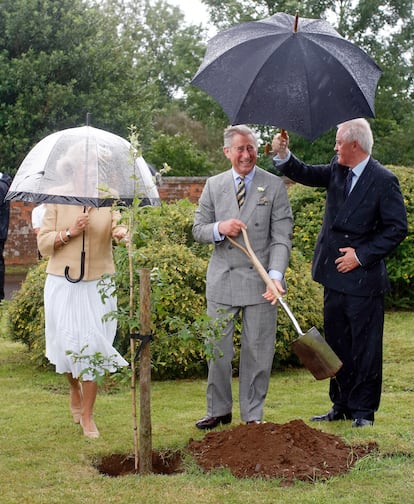 El entonces todavía príncipe Carlos, acompañado por Camila, planta un árbol mientras su oficial de protección policial sostiene un paraguas durante una visita al pueblo de Bromham, en Wiltshire, el 17 de julio de 2007.