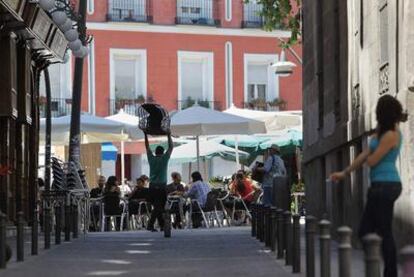 Un camarero lleva más sillas a una terraza situada en la plaza de Tirso de Molina, mientras a la derecha un músico callejero toca el acordeón.