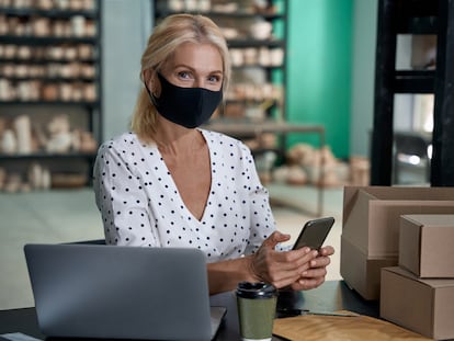 Female business owner wearing protective face mask holding mobile phone and looking at camera while working in her art studio or craft pottery shop with handmade ceramic products - Estar donde estés - Banco Sabadell