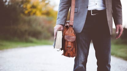 Un hombre camina con un libro en la mano y un bolso tipo bandolera colgado.