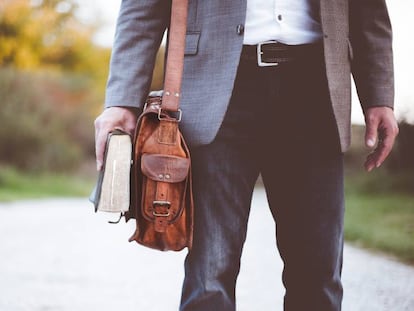 Un hombre camina con un libro en la mano y un bolso tipo bandolera colgado.