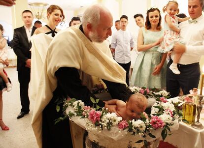 Marius Picu, el párroco, bautiza a la niña con una triple inmersión. Los padres y la hermana de la niña observan con atención mientras la madrina espera con una toalla.