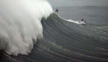 Un surfista trata de mantener el equilibrio bajo la atenta mirada del conductor de una de las motos de agua que acompañan a los deportistas en el agua. Las motos, con las que se ha creado la modalidad del 'tow in' en las que el surfista es arrastrado para entrar en la pared con velocidad, se han convertido en una pieza clave para el surf de olas grandes ya que hacen las veces de socorristas.