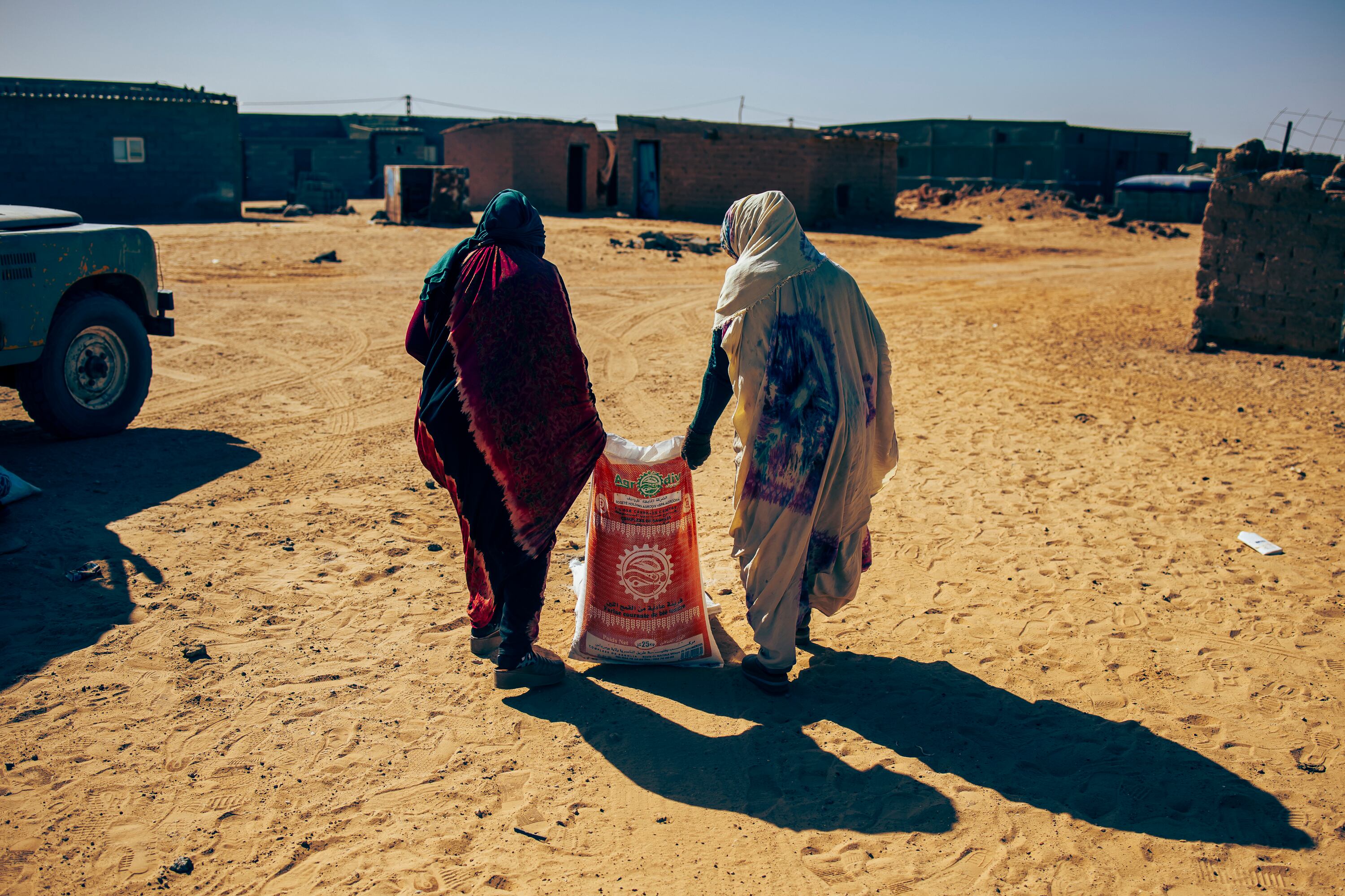 Dos mujeres arrastran un saco de comida hacia su vivienda en el campamento de Auserd. Algunas familias se ponen de acuerdo para recoger juntas su canasta y que no les abran los sacos de comida para dividirlo en raciones. De esta manera, pueden vender el producto más abundante, que es la harina, a otras familias para alimentar a sus animales, las panaderías o pizzerías, y comprar otros alimentos con los ingresos que obtienen. 