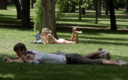 Una mujer lee tumbada en el Parque del Retiro de Madrid, tras dos jóvenes.