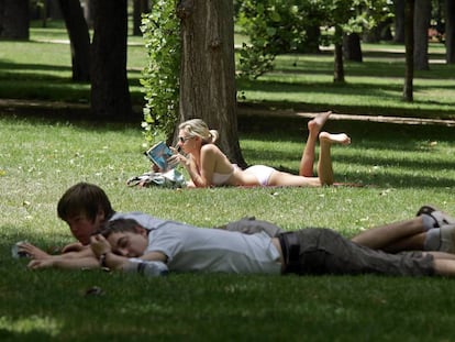 Una mujer lee tumbada en el Parque del Retiro de Madrid, tras dos jóvenes.