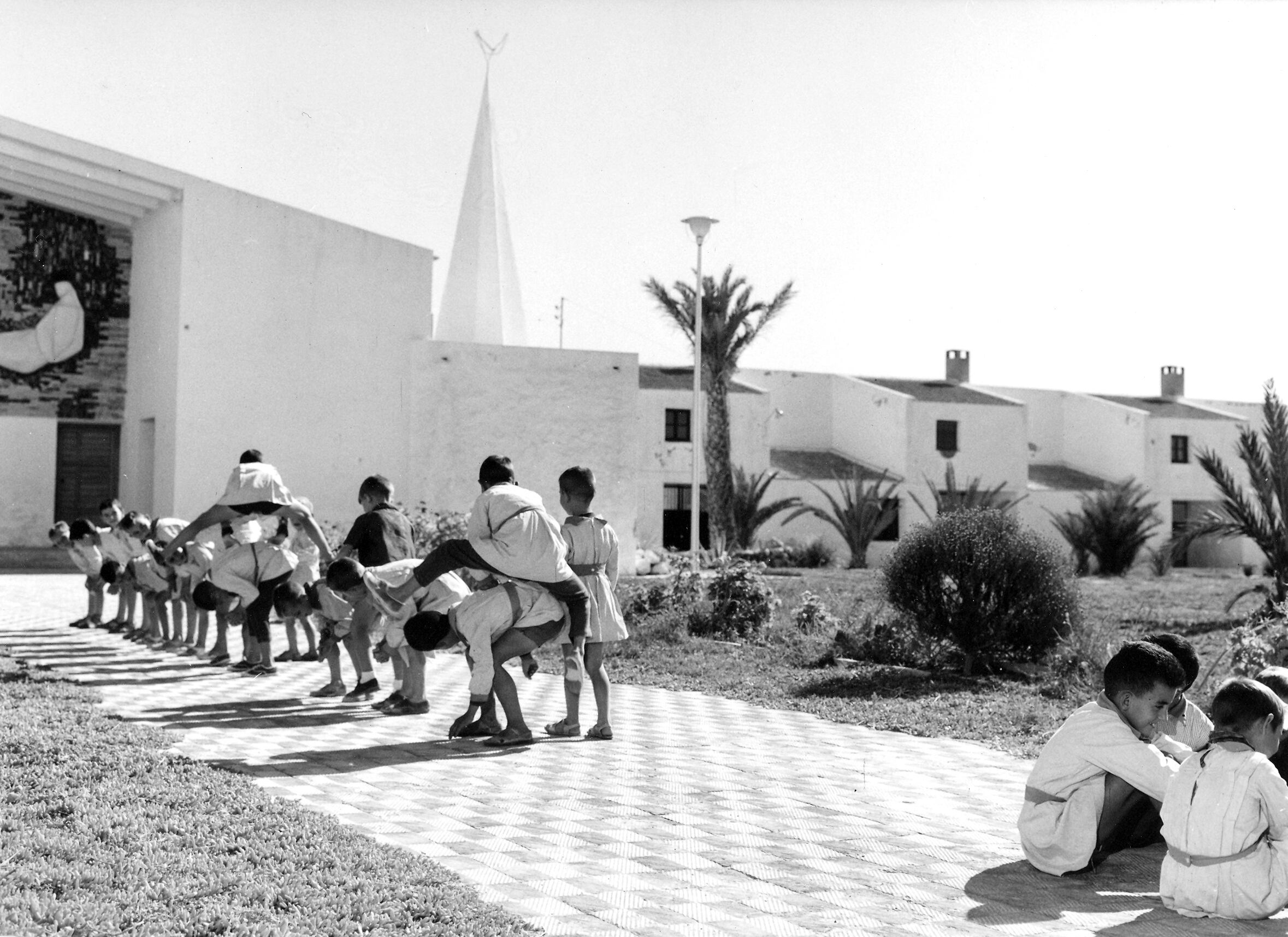 Plaza de la iglesia de El Realengo, Alicante. Hora del recreo escolar.  El mural de la fachada, realizado en ladrillo, teselas cerámicas y piedra, es obra de Manuel Baeza y Adrián Carrillo. Imagen proporcionada por el Ministerio de Agricultura, Pesca y Alimentación, Archivo Central Fondo Instituto Nacional de Colonización. XXV Aniversario, Alicante. Signatura: Hora de recreo.