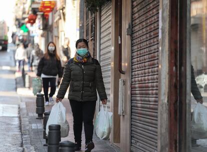 Varias personas caminan protegidas por mascarillas mientras sujetan bolsas de la compra, por una calle del distrito de Usera.