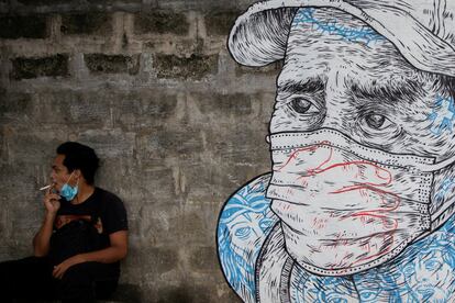 Un joven se fuma un cigarrillo junto a un mural de un hombre con mascarilla, en la ciudad filipina de Quezon.