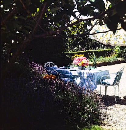 Un rincón del jardín de La Paisible donde se encontraba una mesa de almuerzo cubierta con un mantel de motivos florales acompañada de sillas blancas con cojines azules. Sobre la mesa, una pequeña variedad de cristalería moderna, incluida una jarra alta, rodea un arreglo de rosas en un jarrón plateado.