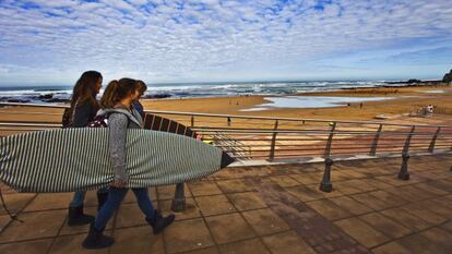 Surferas en la playa de Sopelana, el caser&iacute;o de Landetxo y una fila de copas de vino preparadas para una cata de chacol&iacute;.