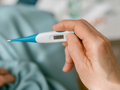 Over-the-shoulder view of an unrecognizable woman looking at the thermometer because she is sick while sitting on the sofa covered with a blanket.